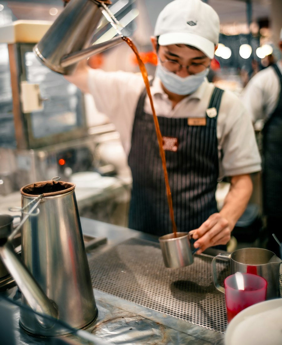 Barista in a cap and mask pulling kopi, pouring it from a height into a metal jug