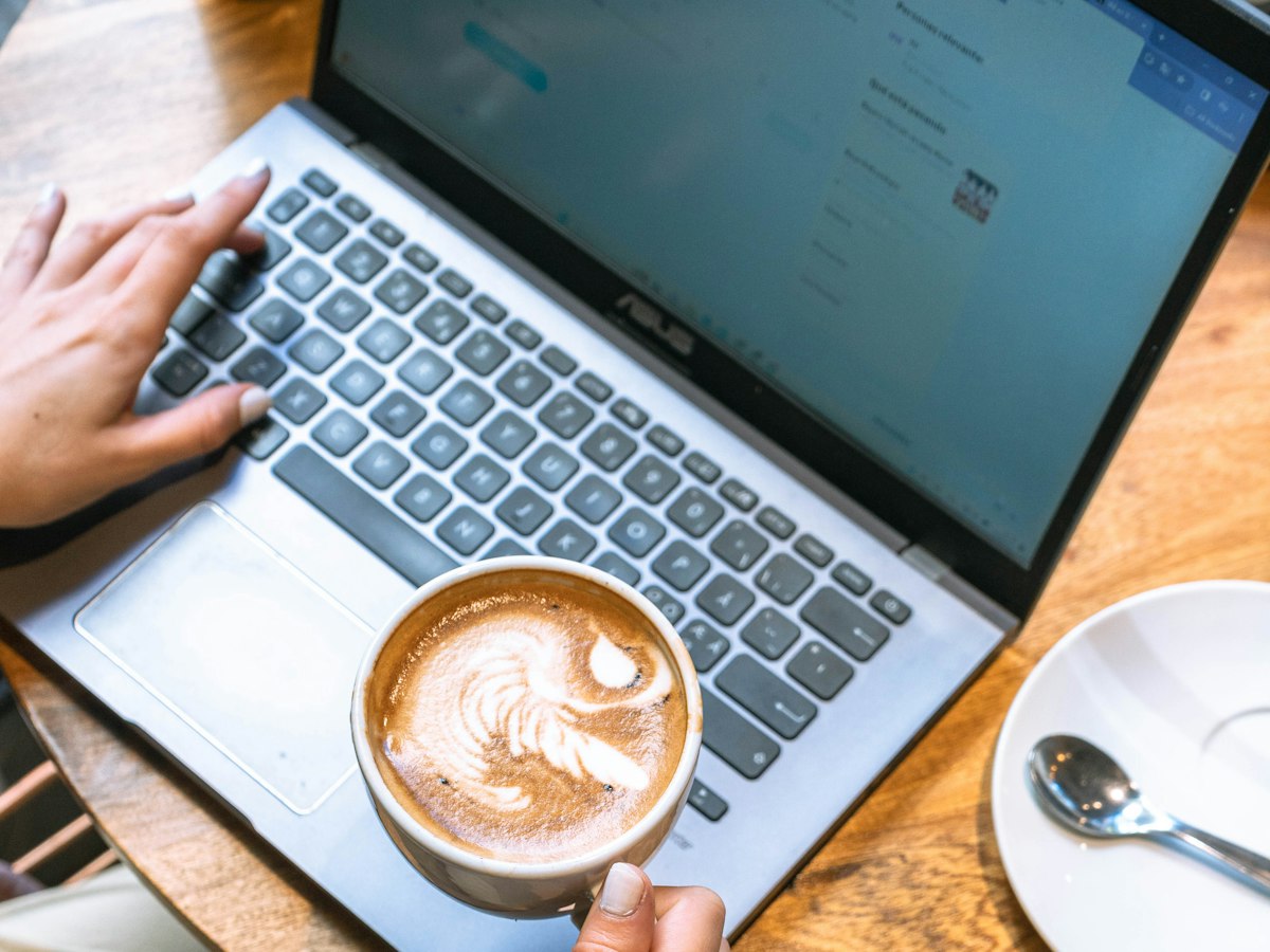 A latte held over an open laptop on a wooden café table